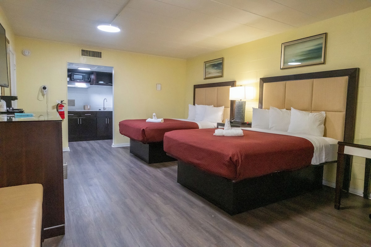 Ein Hotelzimmer mit zwei Betten, einem Schreibtisch, einem Stuhl, einer Mikrowelle und einem Spülbecken in der angrenzenden Küchenzeile.