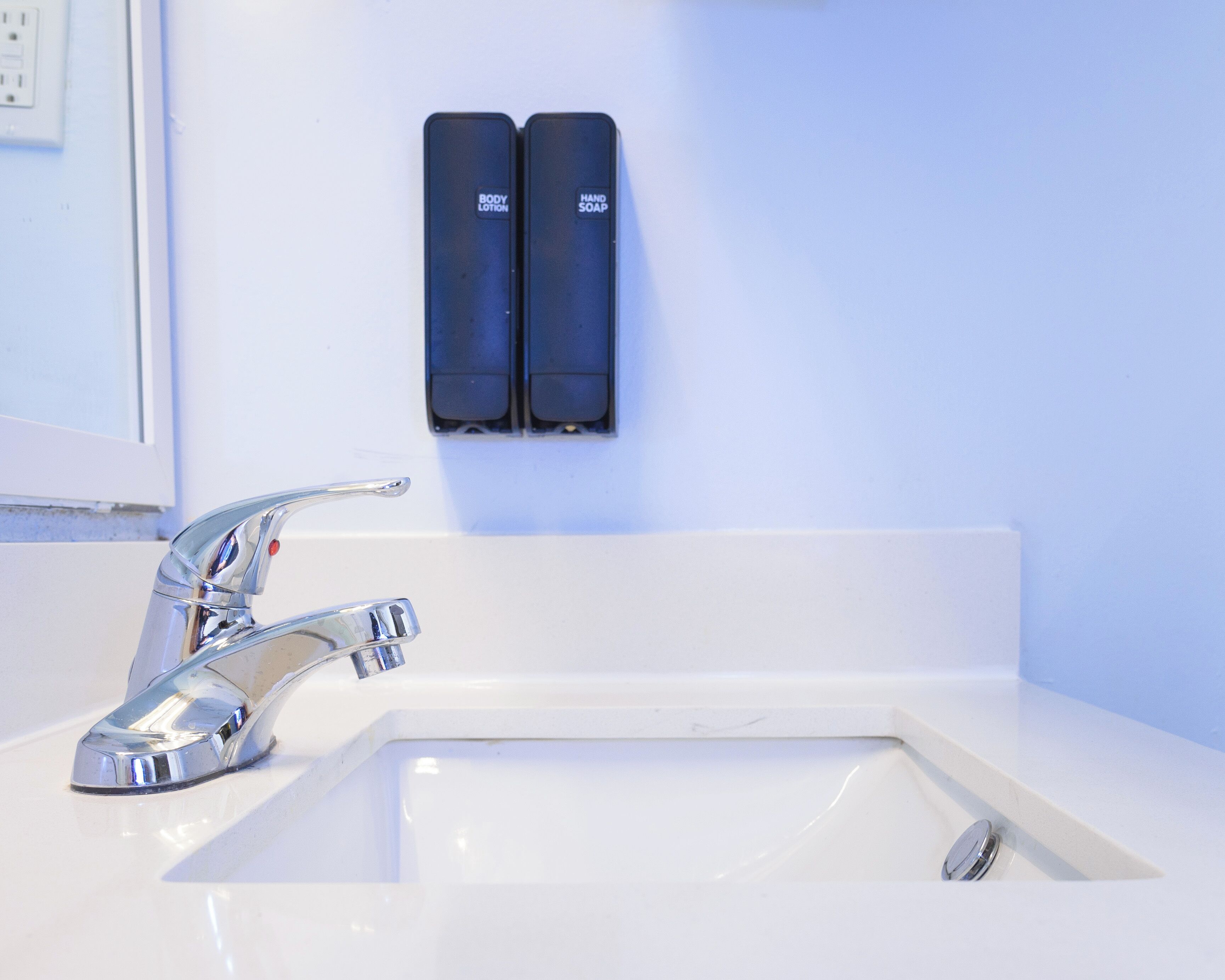 Ein Badezimmer mit einem chromfarbenen Wasserhahn, einem weißen Waschbecken und zwei schwarzen Seifenspendern, an denen „BODY LOTION“ und „HAND SOAP“ befestigt sind.