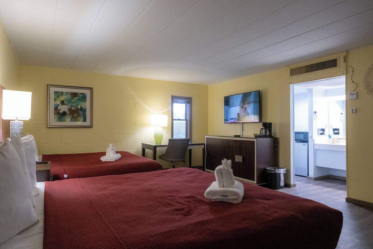 Ein Hotelzimmer mit zwei Betten, einem Schreibtisch, einem Sessel, einem Fernseher und einem durch eine offene Tür einsehbaren Badezimmer.