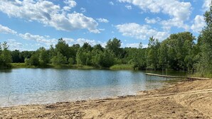 A natural landscape with a lake, sandy shore, and dense trees under a partly cloudy sky.