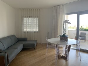 Uma sala de estar moderna com um sofá cinzento, uma mesa de vidro redonda e cadeiras transparentes. Há uma janela com persianas e vista para o verde lá fora.