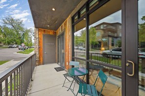 A hotel entrance with a glass door, a small outdoor seating area with a table and chairs, and a view of the street and buildings in the background. - Large & Stylish Glacier National Park Condo! Best Location, Walk Everywhere! (Whitefish)