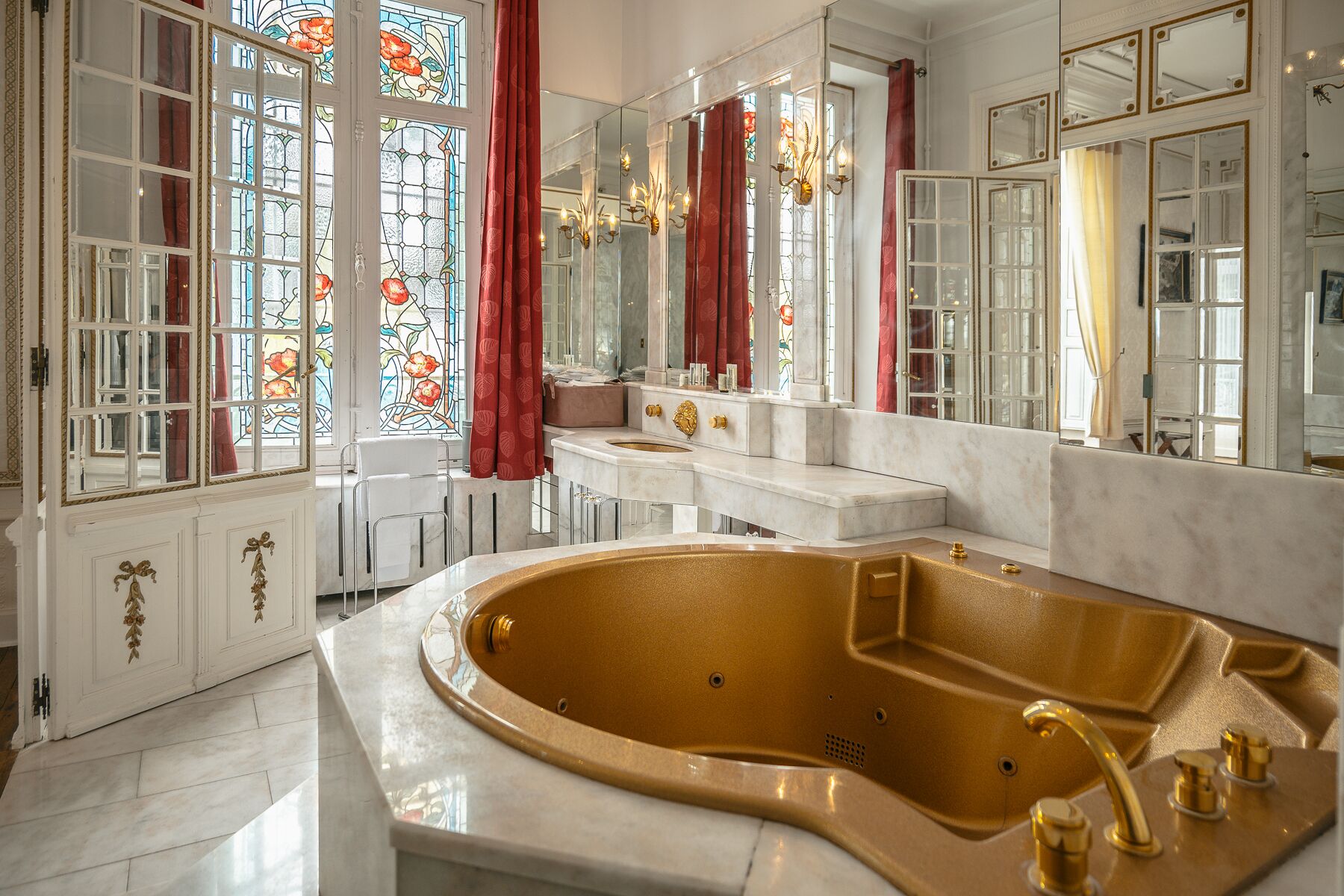 Une salle de bain luxueuse avec une grande baignoire dorée, des comptoirs en marbre blanc et des vitraux.
