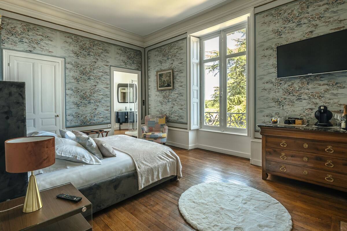 Ein Schlafzimmer mit einem Bett, einem Schminktisch, einem Sessel, einer Lampe, einem Spiegel und einem Fenster mit Blick auf Bäume.