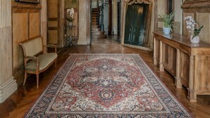 Un amplio pasillo con candelabro, espejos ornamentados, un tapiz grande y una escalera que conduce a otra habitación.