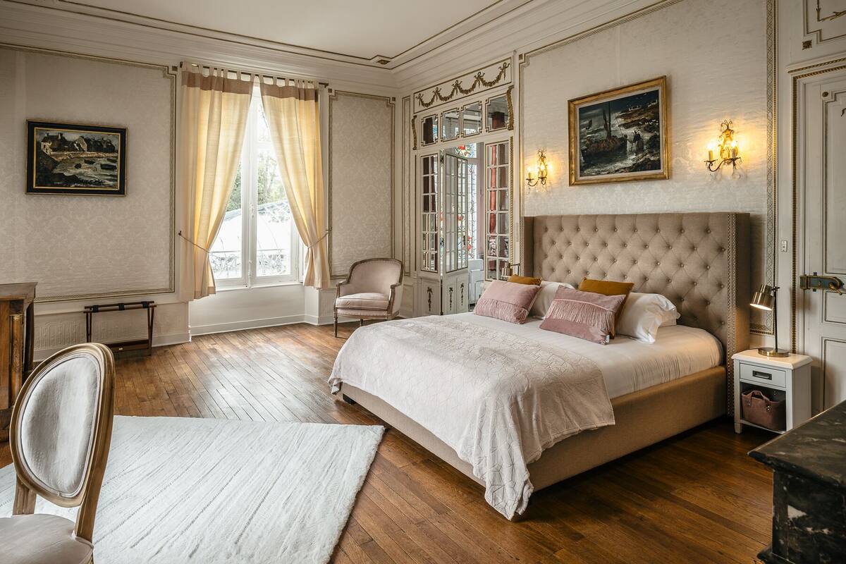 Ein klassisches Schlafzimmer mit einem großen Bett, einem Sessel, einem Schreibtisch und einem Fenster mit Vorhängen.