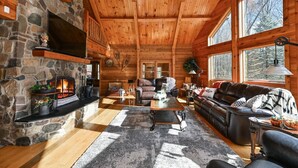 Interior acogedor de una cabaña de madera con chimenea de piedra, muebles de cuero y amplias ventanas con vistas al bosque.