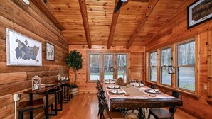 Comedor de una cabaña de madera con una mesa grande dispuesta para varias personas, cuadros en las paredes y ventanas amplias.
