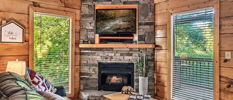 Interior de una cabaña con chimenea de piedra, un televisor montado y una zona de estar acogedora.