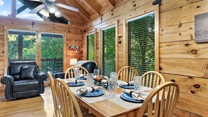 L’intérieur d’un chalet en bois présente une table à manger dressée pour quatre personnes, entourée de chaises en bois, d’un canapé en cuir et d’un ventilateur de plafond.