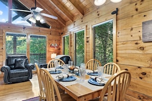 A wooden cabin interior with a dining table set for four, surrounded by wooden chairs, a leather sofa, and a ceiling fan. - Near Dollywood⛰Smokies⛰Hot Tub⛰Pool Table⛰Jacuzzi (Pigeon Forge)