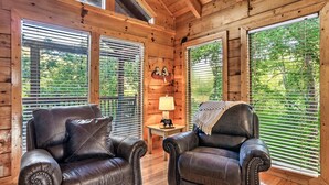 L’intérieur d’un chalet accueillant, avec deux fauteuils en cuir, une petite table avec une lampe et de grandes fenêtres équipées de stores.