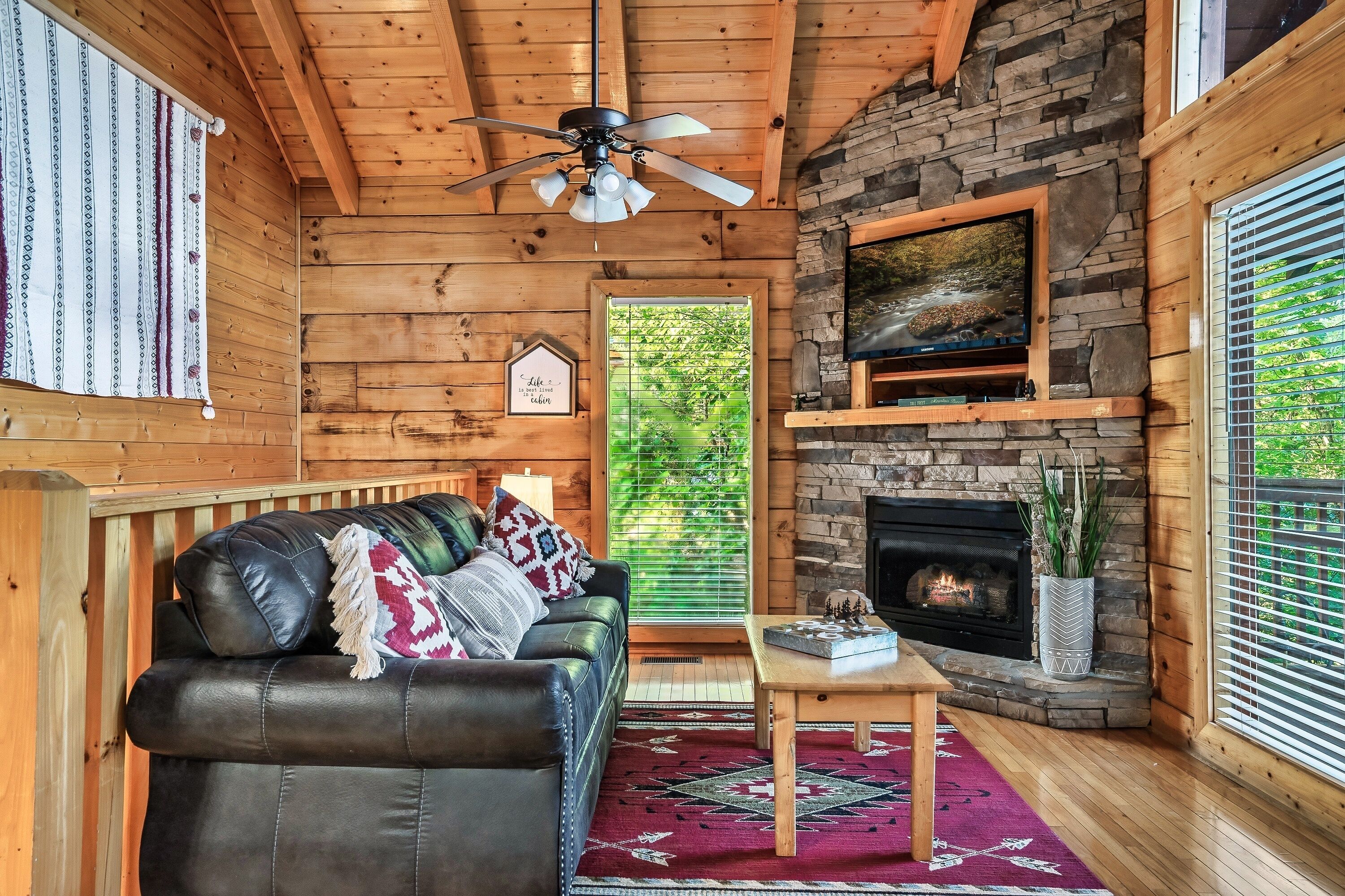 L’intérieur d’une cabane est aménagé avec un canapé en cuir, un ventilateur de plafond, une cheminée en pierre et un téléviseur fixé au mur.