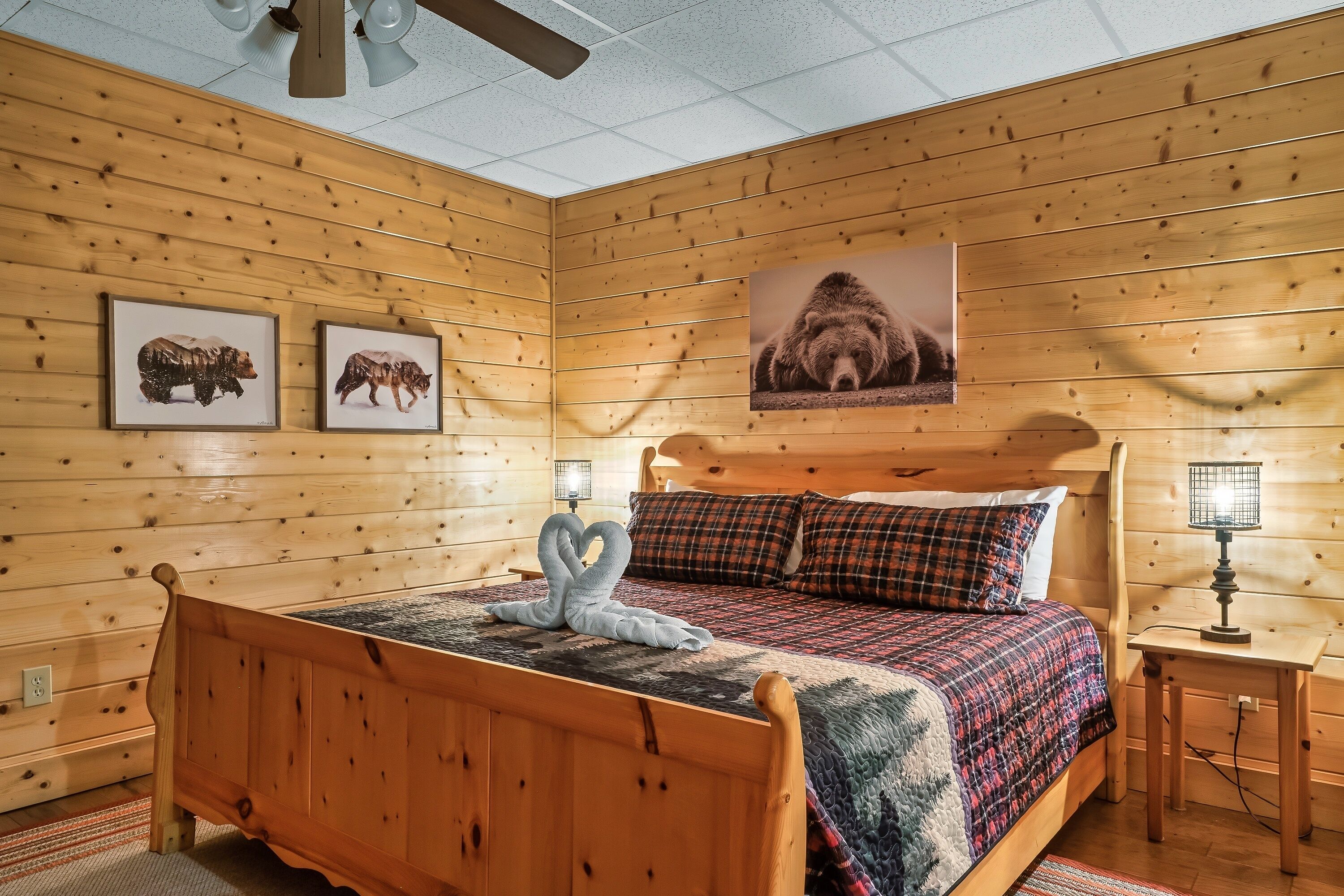 A wooden-paneled room with a bed, two framed animal pictures, a wall-mounted lamp, and a ceiling fan.