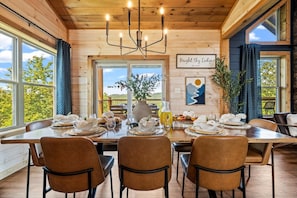 A dining area with a wooden table set for four, surrounded by chairs, with a view of greenery outside.