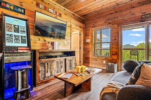 A cozy cabin interior with a wooden interior, a television mounted on the wall, a table with snacks, and a view of the outdoors through the windows.