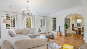 A spacious living room with a large beige sectional sofa, a coffee table, and a chandelier. There are multiple windows with white curtains, a dining area with a table and chairs, and a hallway leading to another room.