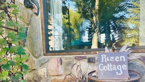 A rustic stone wall with a window reflecting trees, a sign reading "Rizen Cottage," and a garden with plants and rocks.