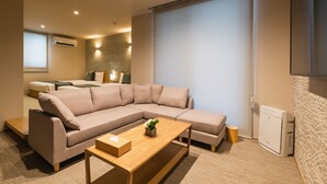 A modern living room with a beige sectional sofa, a wooden coffee table, and a small side table. There is a window with blinds, a wall-mounted air conditioner, and a small cabinet. - GRAND BASE Nagasaki Nakamachi (Nagasaki)