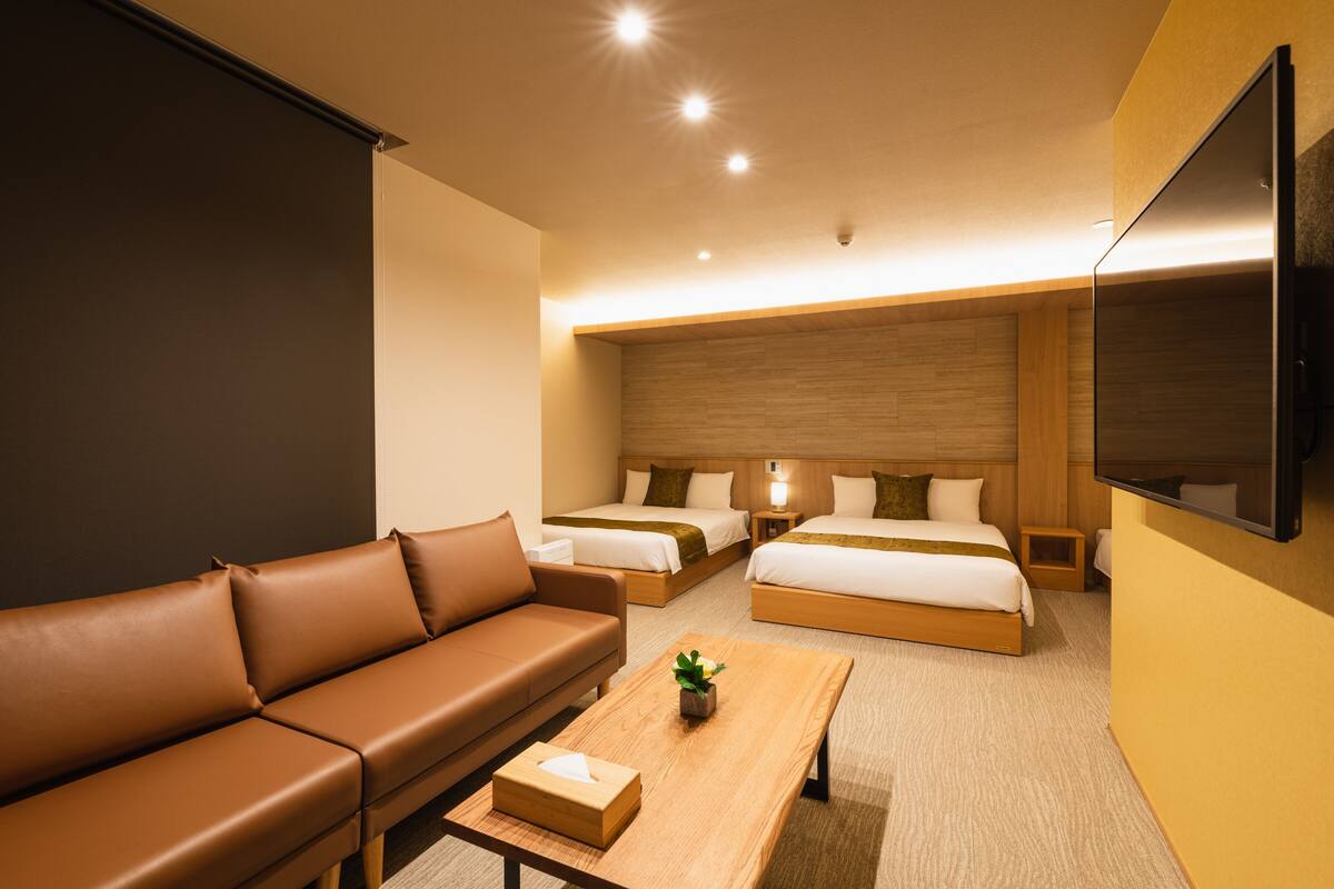 A hotel room with a brown leather sofa, two single beds, a double bed, a wooden coffee table with a plant, and a flat-screen TV mounted on the wall.