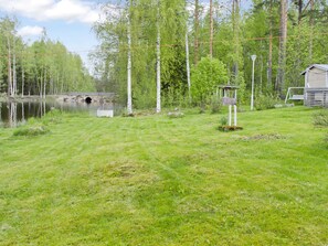A grassy area with a wooden structure, a small bridge over a body of water, and a forested background. - Lietukka by Interhome (Outokumpu)