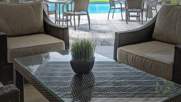 A covered outdoor seating area with wicker furniture and a glass table, overlooking a pool and lush greenery. - Royal Retreat | 2BR • 5 Min Walk to Galleria (Houston)