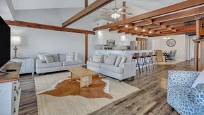 Ein geräumiges Wohnzimmer mit hohen Decken, freiliegenden Holzbalken, einer zentralen Sitzecke mit zwei Sofas, einem Couchtisch und einem Essbereich mit Tisch und Stühlen.