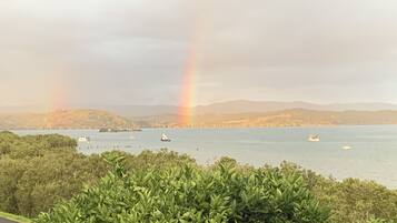Un paysage côtier avec un arc-en-ciel se déployant au-dessus d’une étendue d’eau, des bateaux et des collines en arrière-plan.