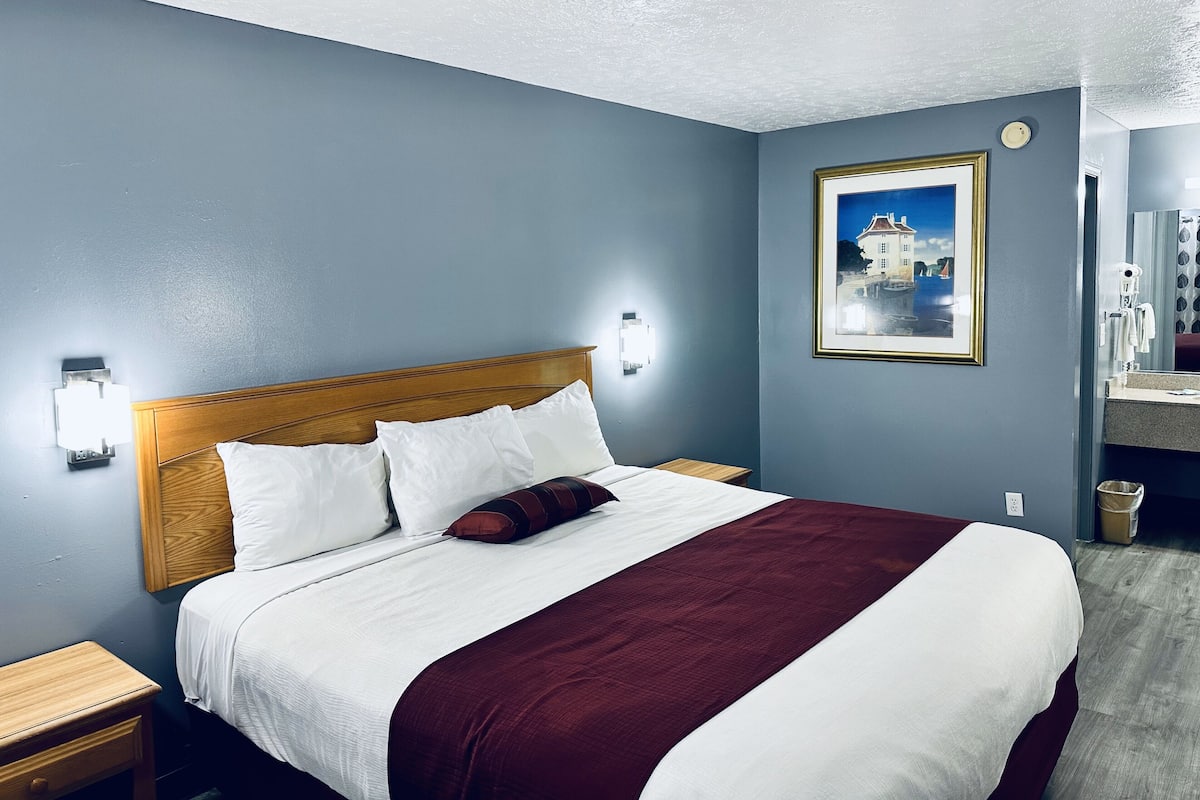 A neatly made bed with white and maroon bedding, a wooden headboard, and a nightstand with a lamp.