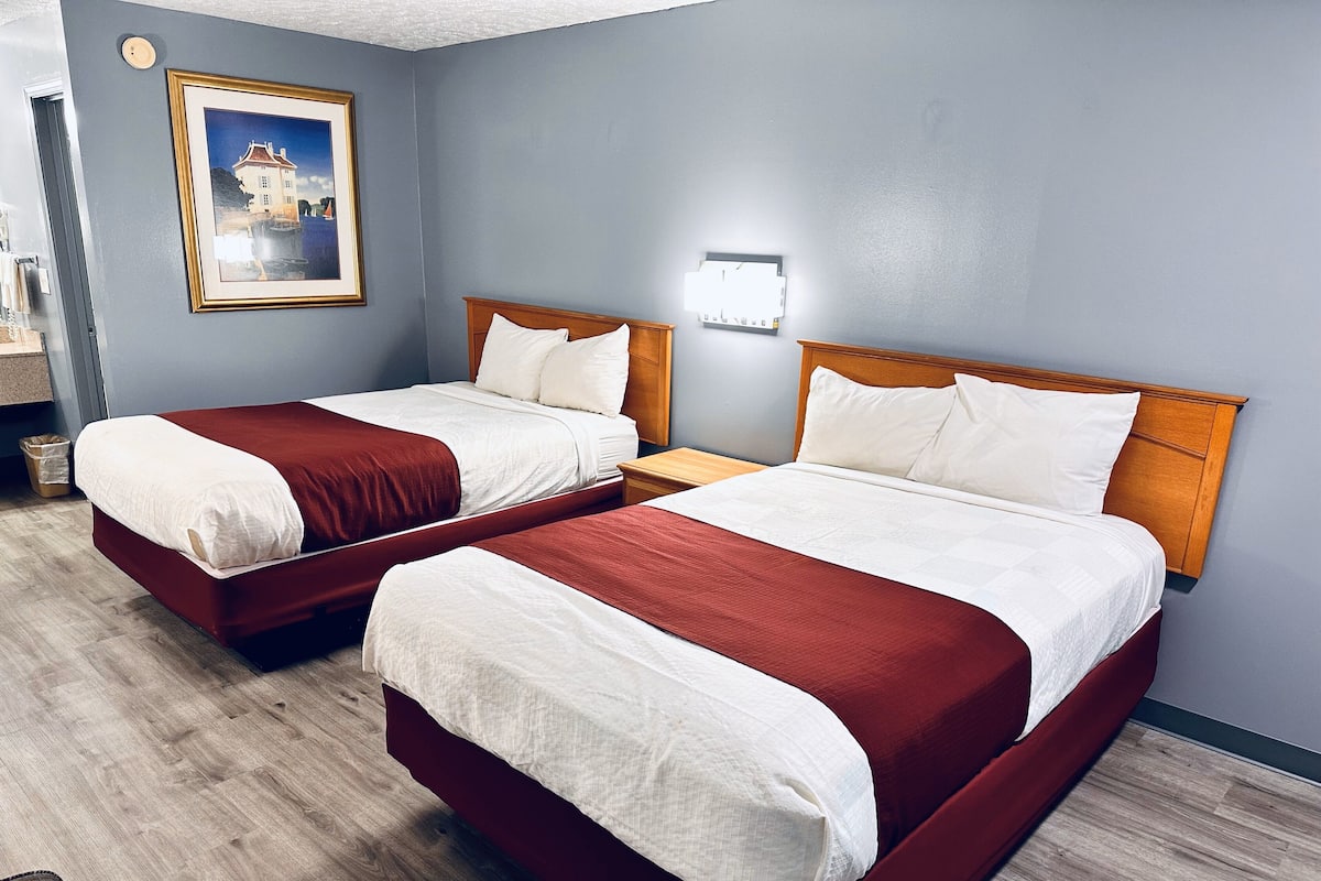 A hotel room with two beds, a painting on the wall, and a nightstand with a lamp.
