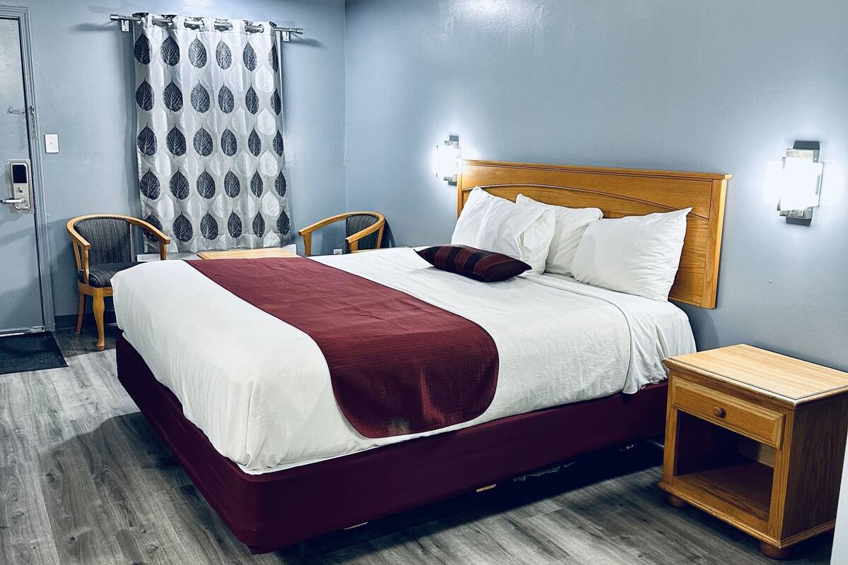 A hotel room with a large bed, a wooden nightstand, a chair, and a wall-mounted light fixture.