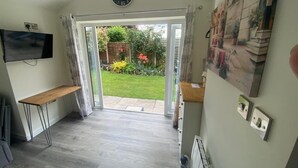 A room with a wooden floor, a flat-screen TV mounted on the wall, a large clock, and a sliding glass door leading to a garden. - Rose View - Modern detached bungalow (Sale)