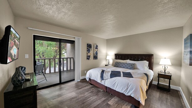 A bedroom with a large bed, a nightstand with a lamp, a chair, a TV, and a sliding glass door leading to a balcony. - Heart of Scottsdale (Scottsdale)