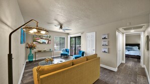 A spacious living area with a sofa, two armchairs, a coffee table, and a TV. There is a ceiling fan, a large window, and a door leading to a bedroom. - Heart of Scottsdale (Scottsdale)