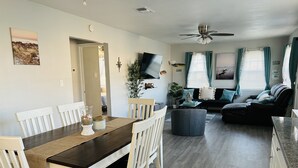Een eetgedeelte met een houten tafel en witte stoelen, een woonkamer met een bank en een televisie, en een plafondventilator.