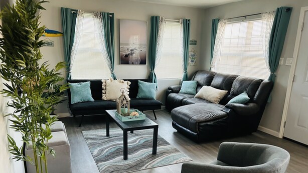 Een moderne woonkamer met een zwarte leren bank, een grijze fauteuil en een salontafel. Er zijn twee ramen met gordijnen, een bloempot en een ingelijst beeld op de muur.