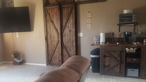 A room with a large sliding barn door, a flat-screen TV mounted on the wall, a wooden bench, a small trash bin, and a kitchenette area with a toaster oven and coffee maker. - COMFY  2 bedroom 1 bath, living room, mini kitchen with 70" smart TV & Grill (Montrose)