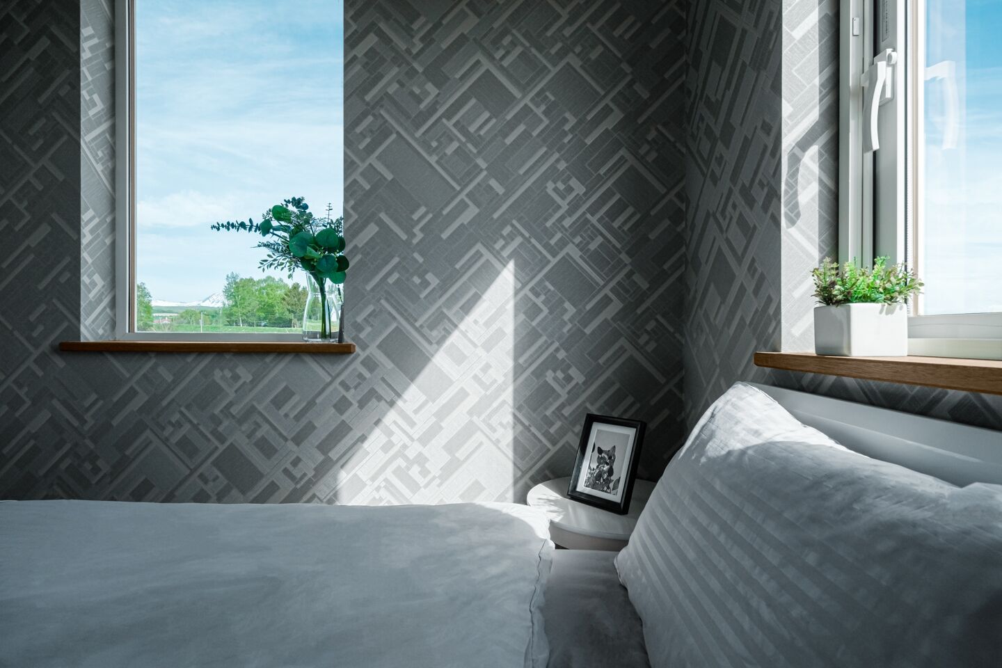 A modern bedroom with a large window, a bedside plant, and a framed picture on the nightstand.