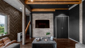 A modern living room with a herringbone-patterned wall, a large flat-screen TV mounted on the wall, and a wooden staircase leading to another level. - THE LANDSCAPE (Kamifurano)