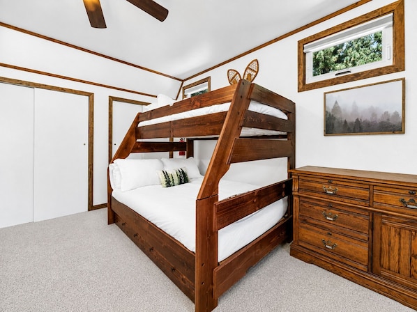 Una cama litera con estructura de madera, ropa de cama blanca, un aparador con cajones y cuadros enmarcados en la pared.