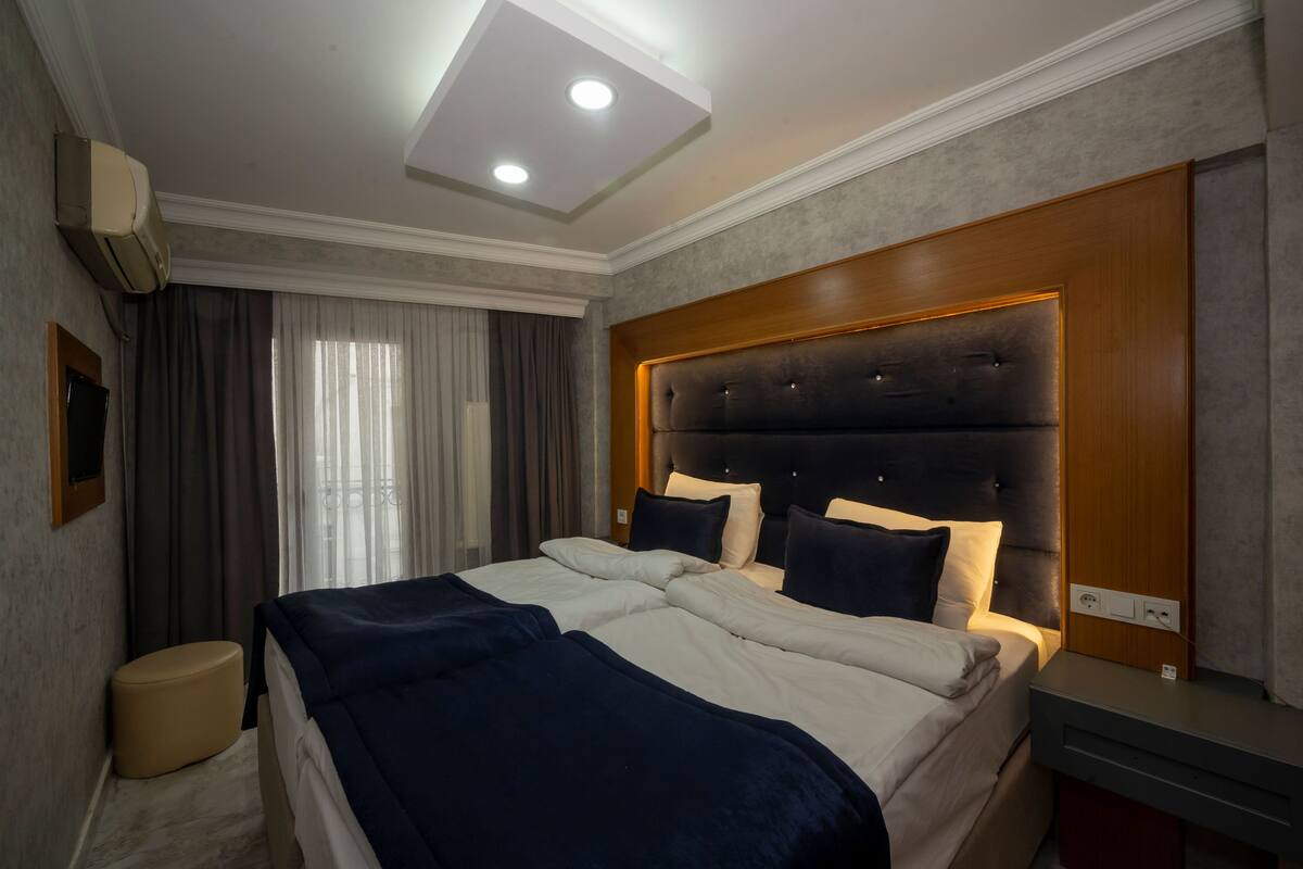 A hotel room with a large bed, a wooden headboard, a bedside table, a wall-mounted air conditioner, and a window with curtains.