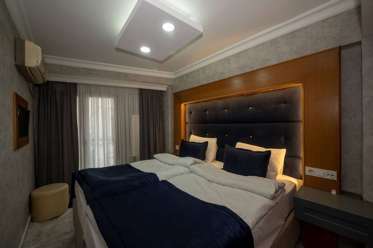A hotel room with a large bed, a headboard, a bedside table, a TV, and a window with curtains.