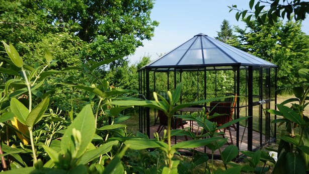 A glass gazebo with a metal frame, surrounded by lush greenery and trees. - Apartment "summer" - Apartments Baltic - Kate (Schmatzin)