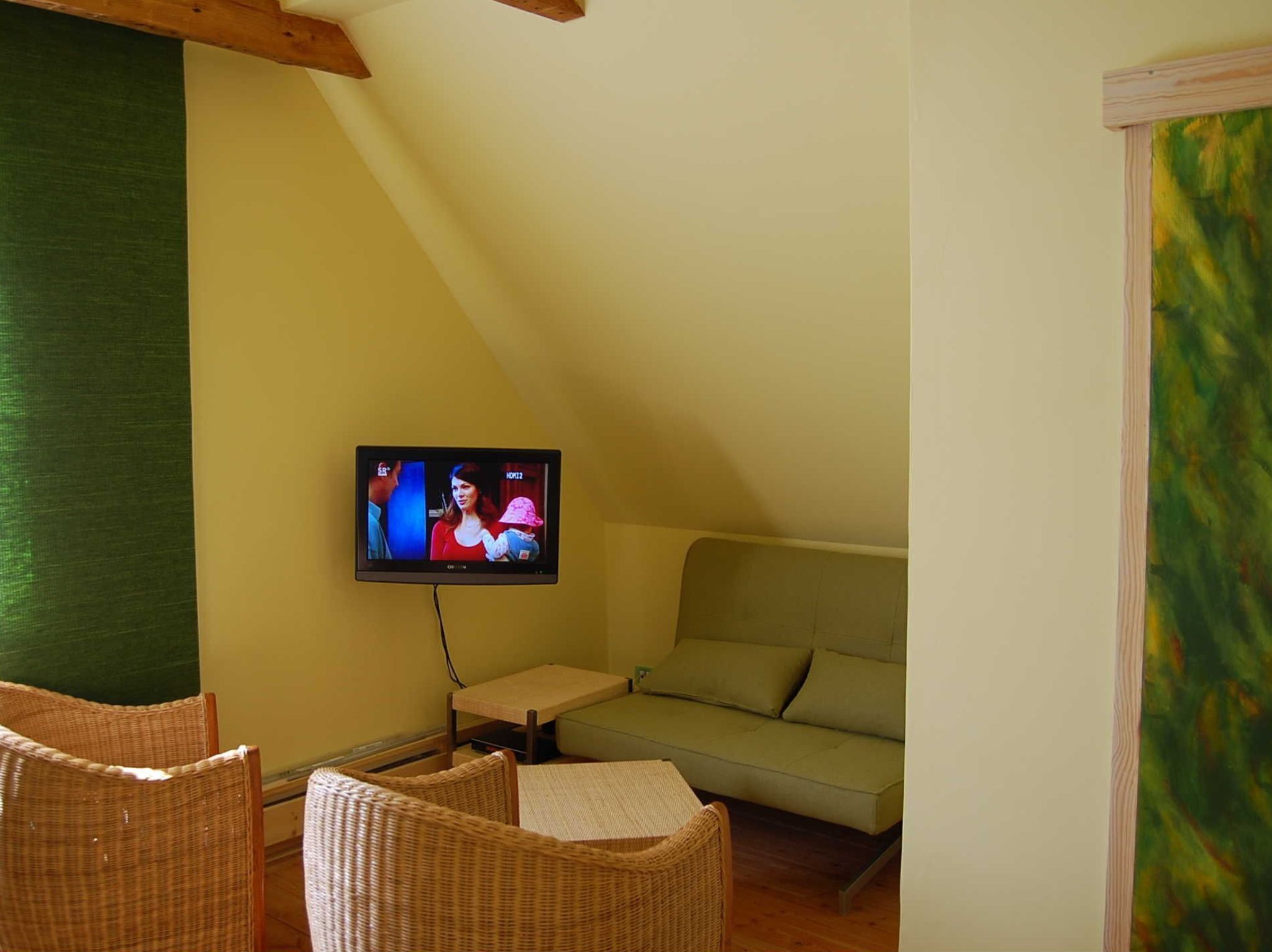 A cozy room with a television mounted on the wall, a green sofa, wicker chairs, and a small table.