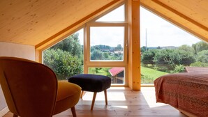 Een slaapkamer met een groot raam, houten vloer, een bed, een gele stoel en een donkere voetenbank.