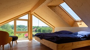 Een kamer met een schuine kap, een bed met een donkere deken, houten vloeren en grote ramen met uitzicht op groen en gebouwen.
