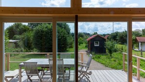 Een houten terras met een tafel en stoelen, een kleine schuur achtergrond en uitzicht op een gazon en bomen.