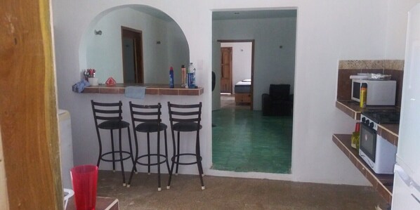 A kitchen with a microwave, stove, and a small refrigerator. There are three black chairs and a red cup on the counter.