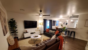 Uma sala de estar moderna com um sofá de couro, um móvel de entretenimento branco, uma TV de tela plana e uma área de jantar com bancada de cozinha.