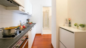 Cocina estrecha con estufa, ollas y fregadero. El suelo es de madera y hay una puerta que conduce a un baño.